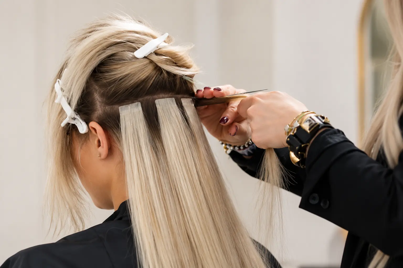 EXTENSIONS DE CHEVEUX 1 MYLAAEVANS Coiffeuse posant des extensions à bandes adhésives sur des cheveux blonds